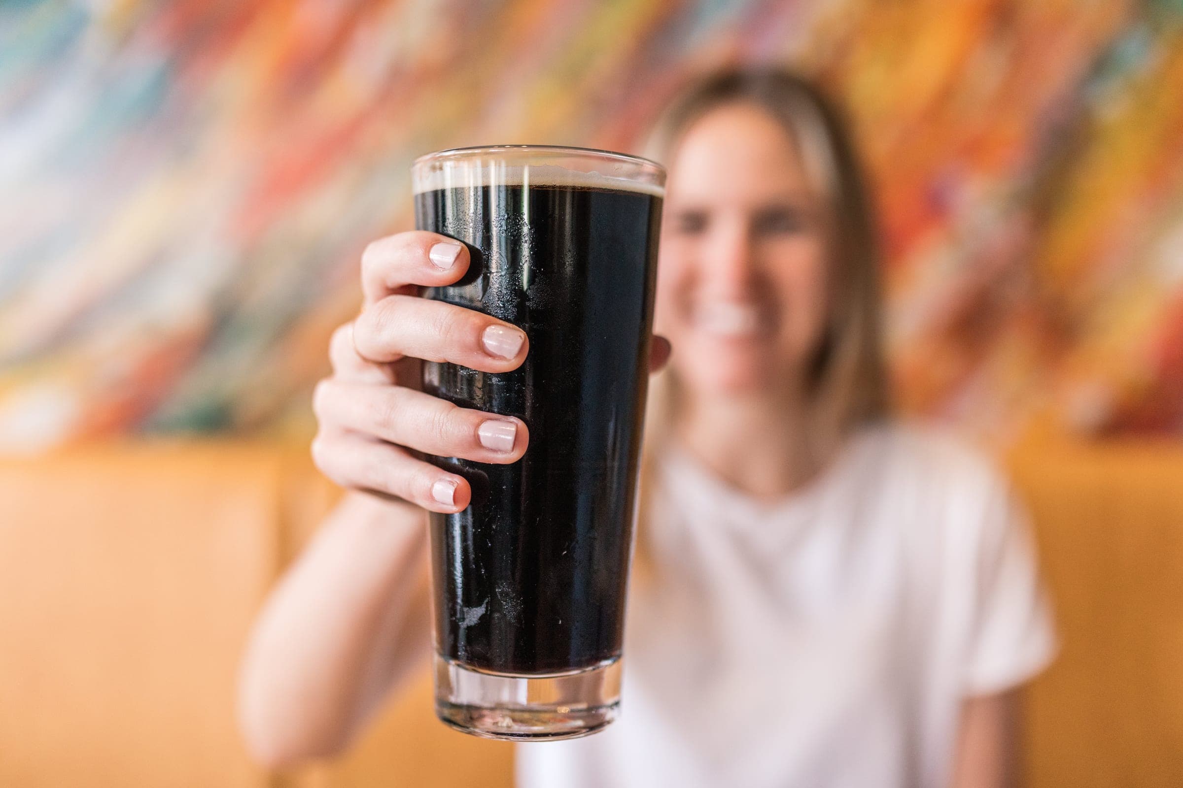 A guest raising a pint of dark craft beer against a colorful mural