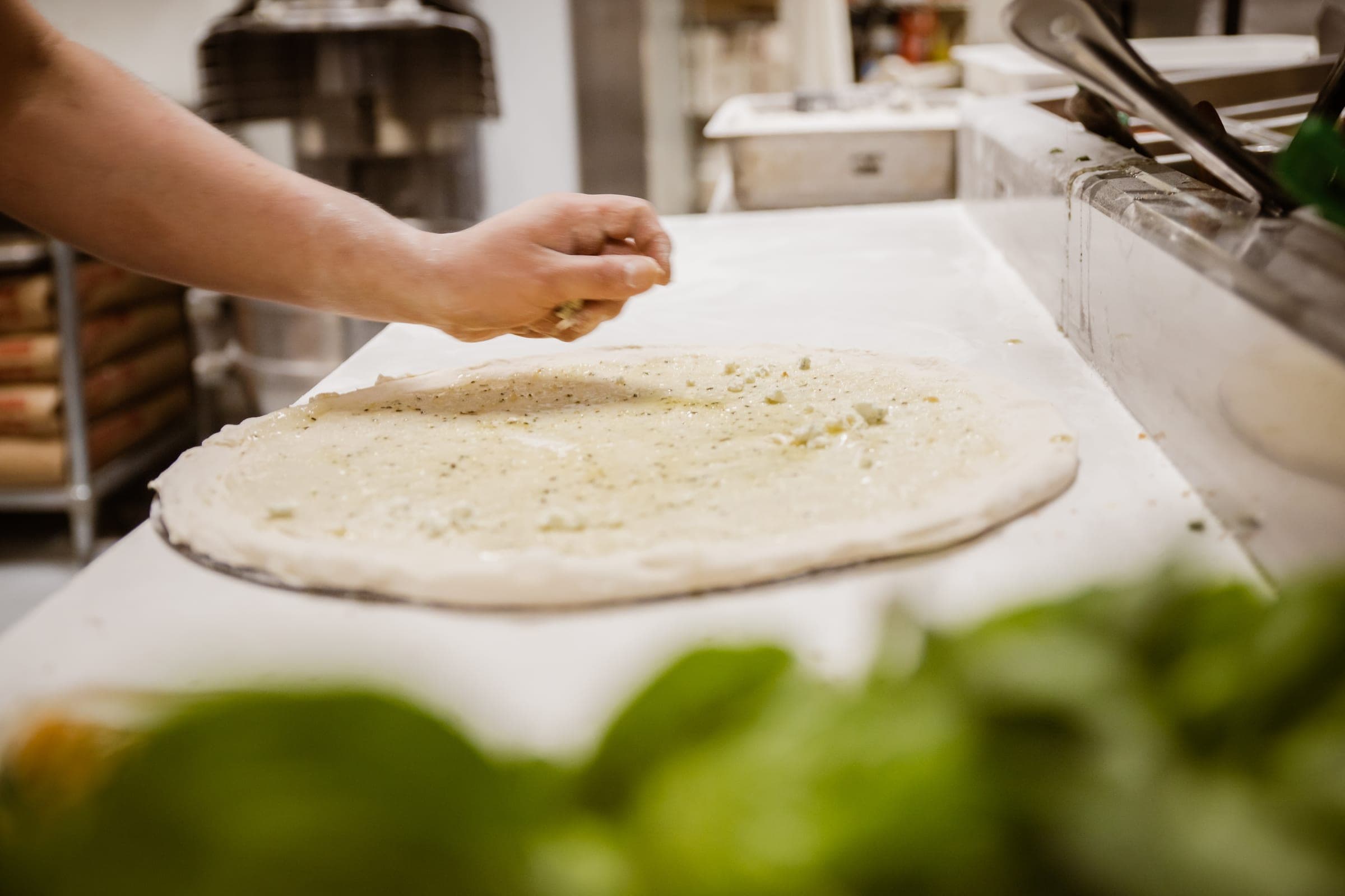 A hand seasoning a freshly stretched dough base on the prep counter on the counter
